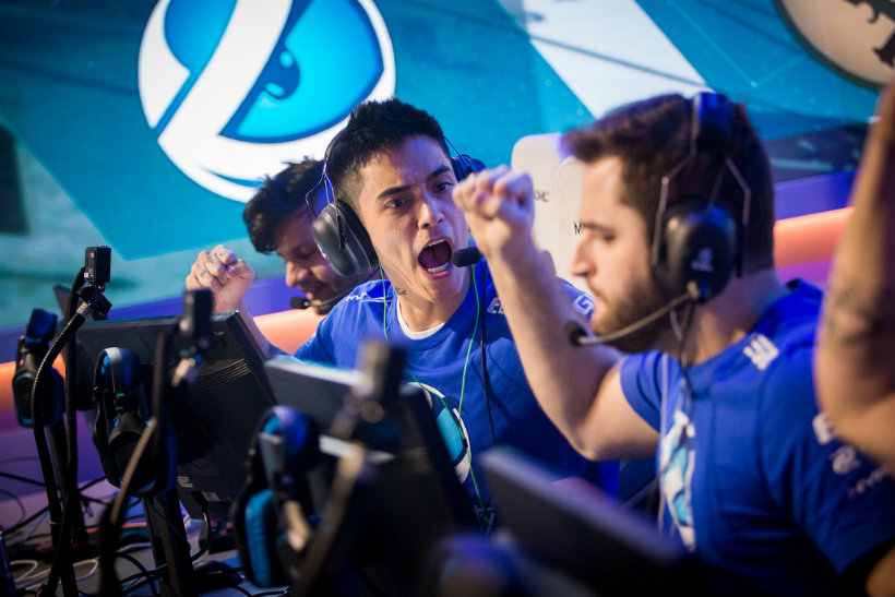 Players wearing blue jerseys with hands in the air celebrate a win at a gaming tournament by their computers